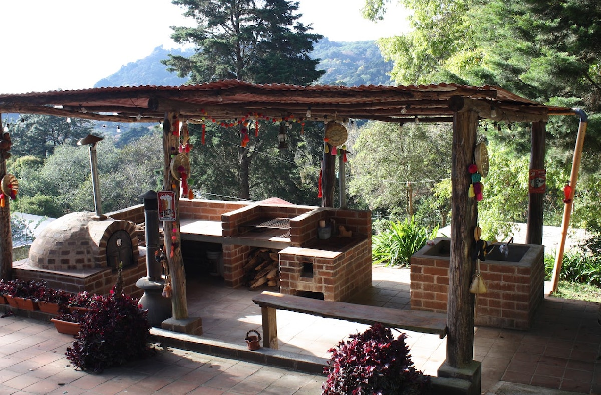 An outdoor cooking area features brick structures for grilling and a traditional pizza oven, surrounded by greenery. Colorful decorations hang from the wood roof, and a wooden bench is positioned for seating. Scenic views of distant hills are visible beyond the space.