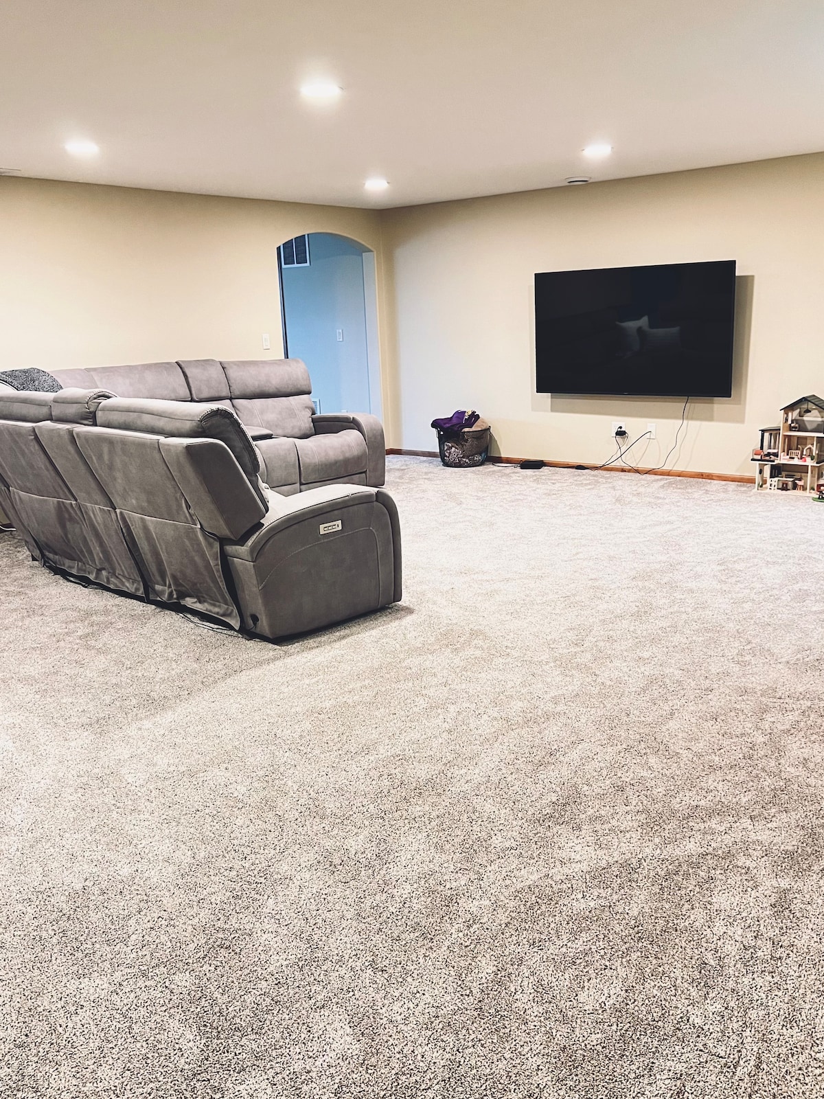 A spacious living area features plush gray carpet and a comfortable reclining sofa. A large flat-screen television is mounted on the wall, providing an engaging focal point. Soft, overhead lighting enhances the feeling of openness, while minimal decor keeps the space uncluttered.