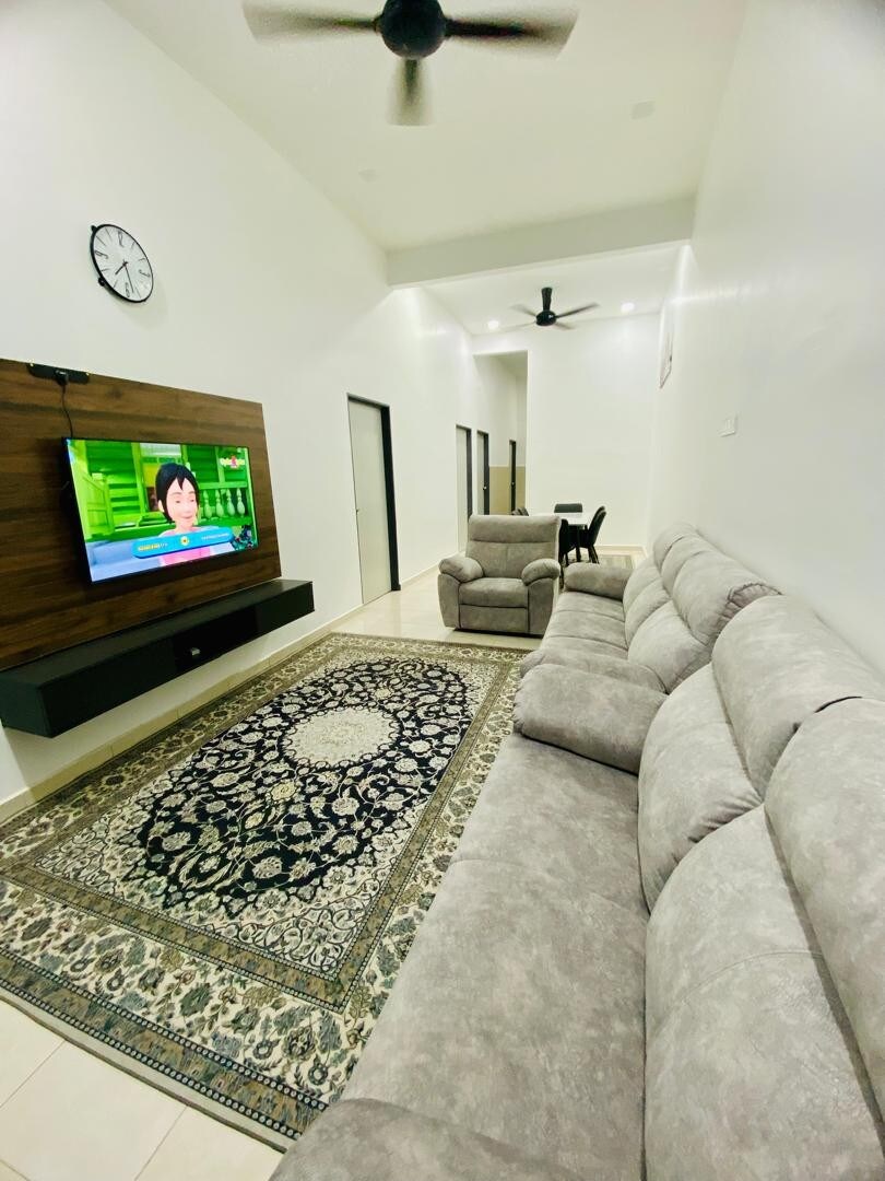 A spacious living area features a neutral-toned sectional sofa positioned near a wall-mounted television. A decorative area rug adds texture to the tiled floor. The room is illuminated by ceiling fans and has a calm, contemporary design with open access to other spaces.
