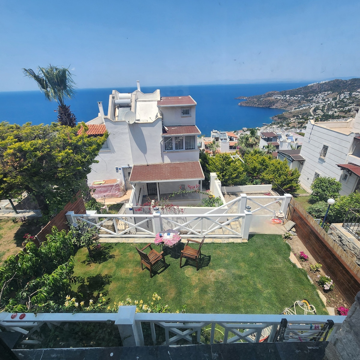 A panoramic view of a well-maintained garden with green grass is framed by white fencing. Two wooden chairs are placed on the lawn, surrounded by blooming flowers. The background features a coastal landscape and a glimpse of the blue sea under a clear sky.