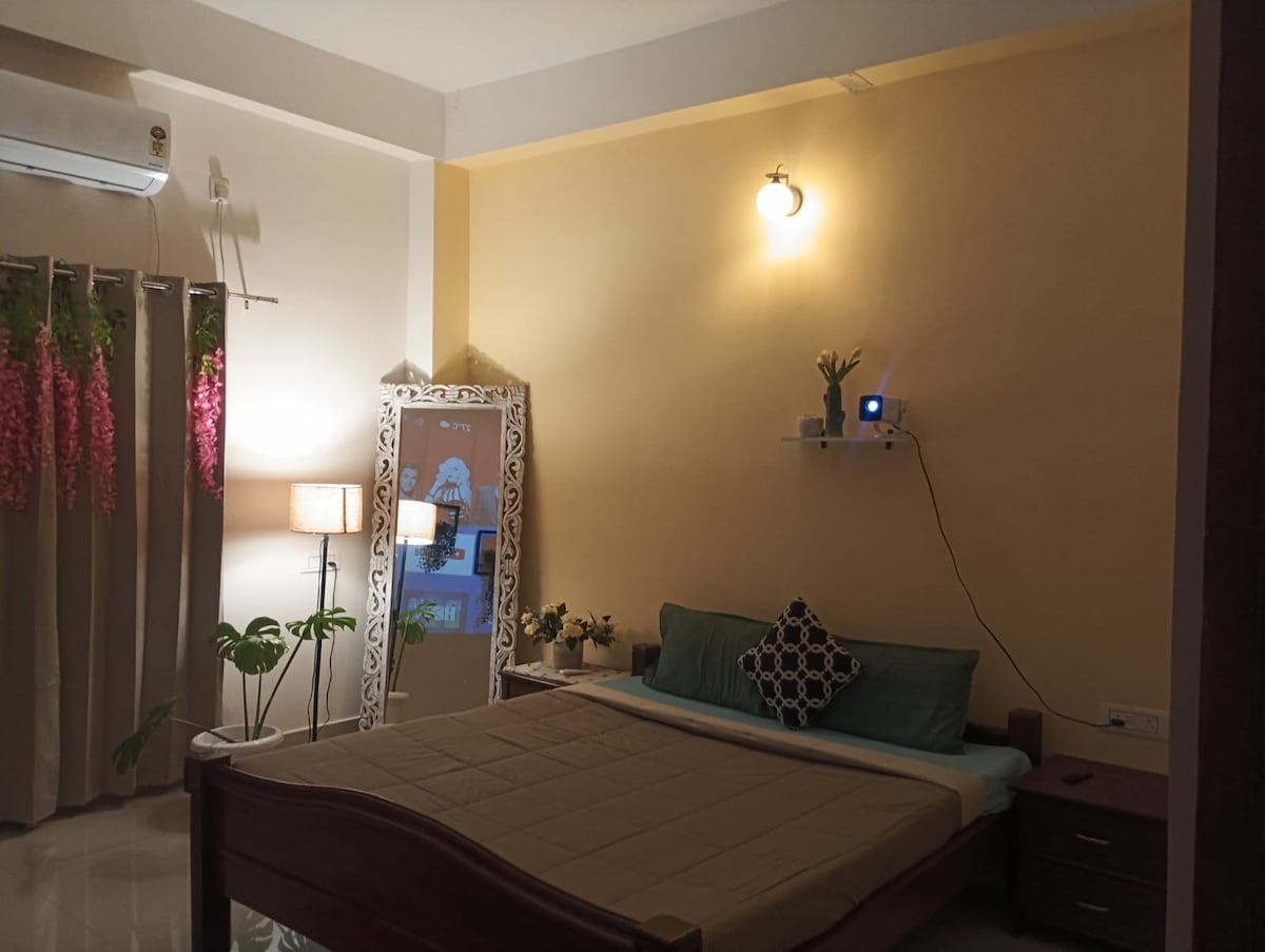 A serene bedroom is equipped with a spacious bed featuring a neutral-colored blanket. A full-length mirror and bedside tables are present. Soft lighting is provided by lamps, with decorative plants enhancing the ambiance. Curtains with floral accents frame a window, allowing glimpses of greenery outside.