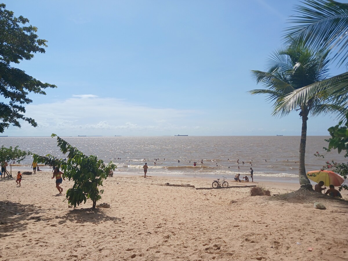 A sandy beach scene is filled with people enjoying the water. Small trees provide shade, while a few bicycles are casually placed nearby. In the distance, ships can be seen on the horizon, under a clear sky, with gentle waves lapping the shore.