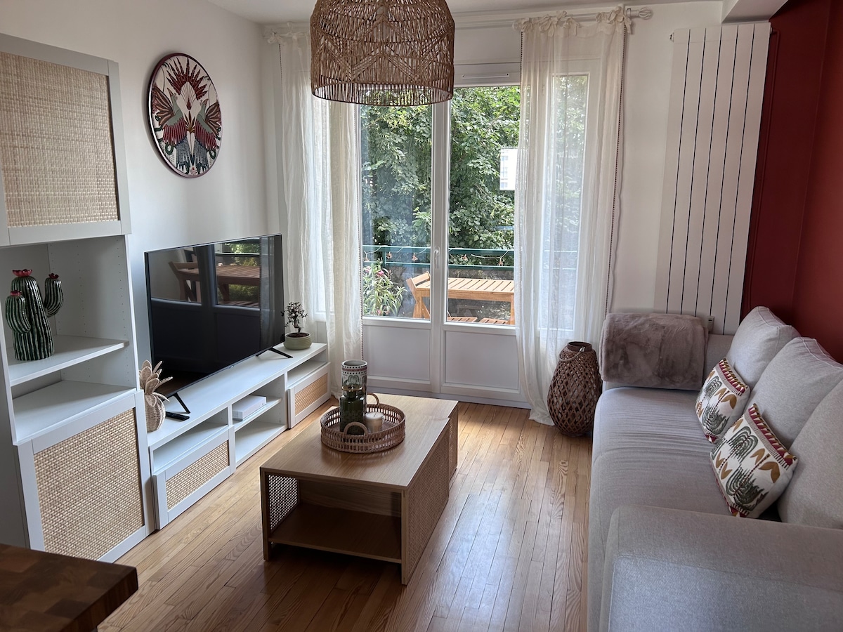 A living area features a light sofa against a vibrant red wall, accompanied by a wooden coffee table. A television is positioned on a white cabinet, and large windows allow natural light to fill the space, providing a view of the greenery outside.