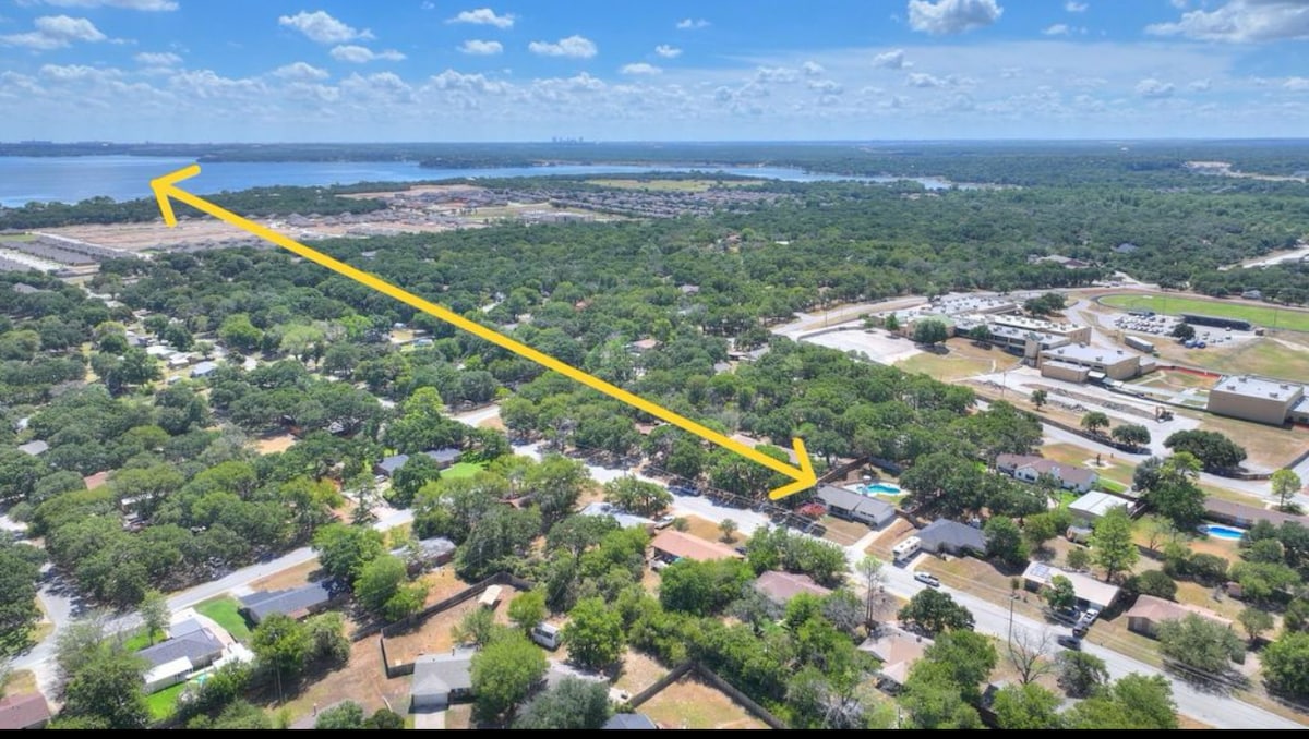 An aerial view captures the surrounding landscape featuring lush greenery and tree-lined neighborhoods. In the distance, Eagle Mountain Lake glimmers under a blue sky dotted with clouds. The image highlights serene residential areas and the proximity of the water.