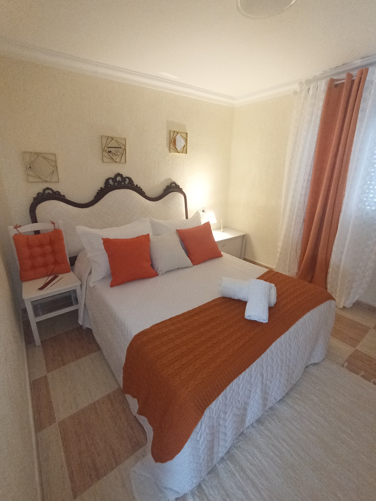 A cozy bedroom is presented with a spacious bed dressed in white linens and an orange throw. Decorative pillows in coordinating colors add a pop of vibrancy. A bedside table and lamp are situated beside the bed, with light filtering through sheer curtains at the window.