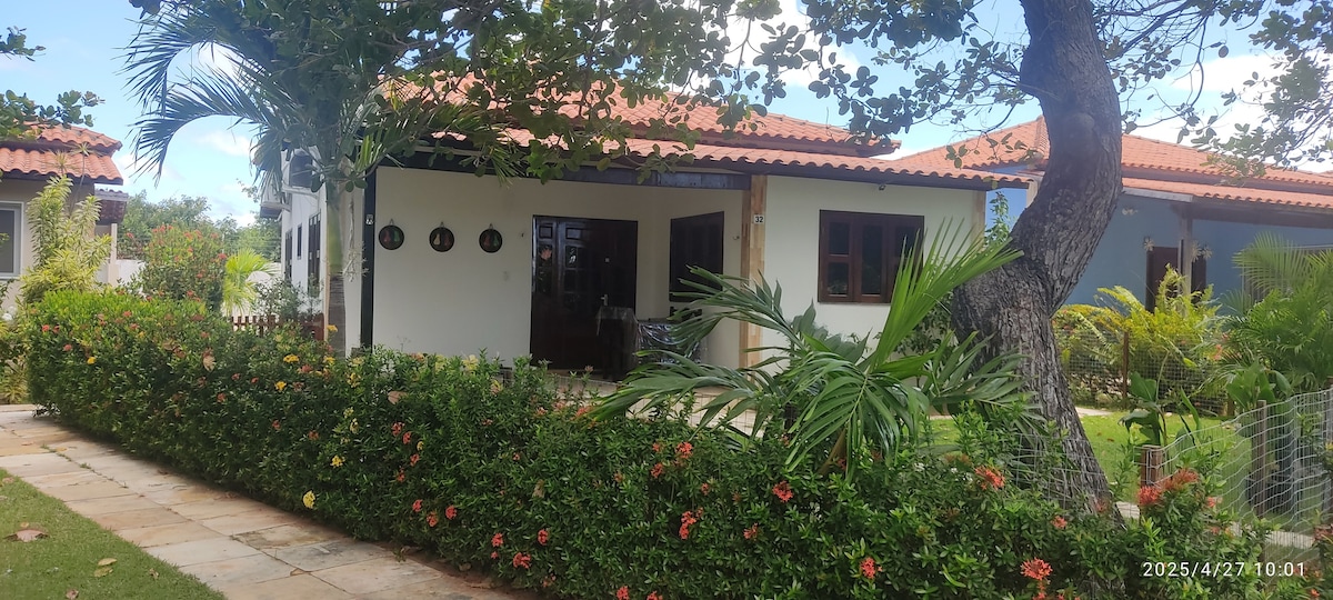 A charming exterior view of a house is presented, showcasing a porch with wooden beams and an inviting entrance. Lush greenery and colorful flowers create a vibrant landscape, while palm leaves gently sway in the breeze beside the pathway made up of stone tiles.