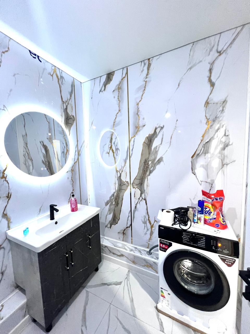A modern bathroom features elegant marble walls with golden veins. A round mirror is positioned above a sleek vanity with a sink. A washing machine and various laundry supplies are placed nearby, enhancing the functional aspect of the space.