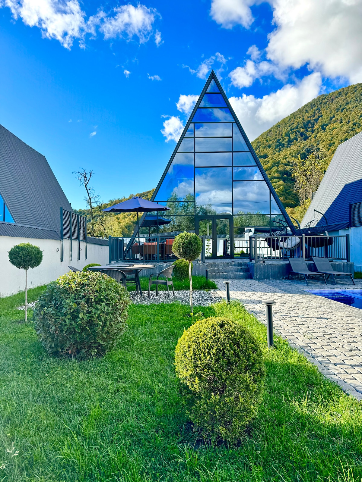 A striking triangular structure with extensive glass panels highlights the contemporary architecture. Lush green grass and neatly trimmed shrubs surround the property, while a swimming pool reflects the sky. The backdrop features a picturesque mountain, enhancing the outdoor space.