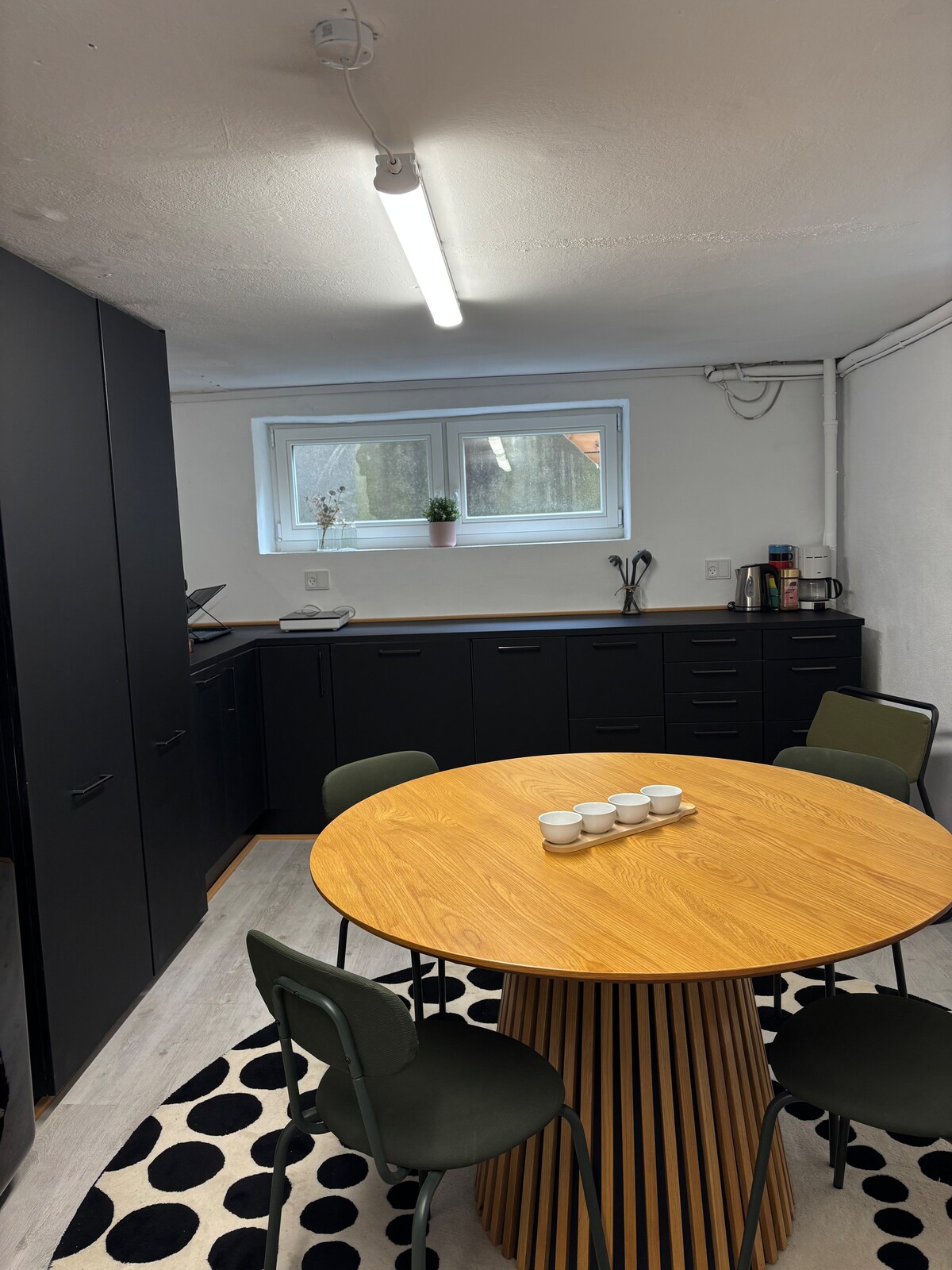 The kitchen area features a round wooden table surrounded by four green chairs. Black cabinetry lines the walls, providing ample storage. A window allows natural light to enter, while a small plant and kitchen appliances are visible on the counter.