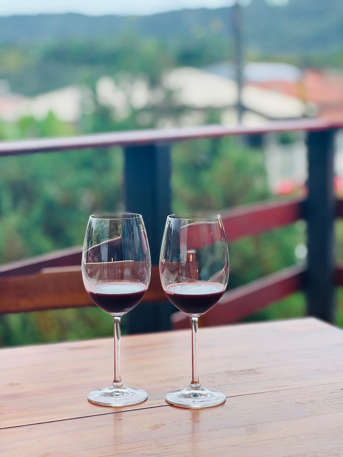 Two wine glasses filled with red wine are positioned on a wooden table. A blurred green landscape and distant buildings are visible in the background, adding depth to the scene.