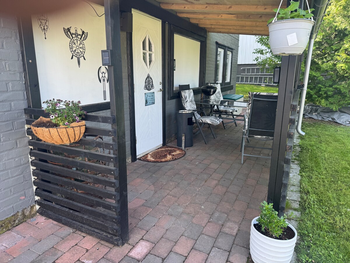 An outdoor seating area is presented, featuring a table and several chairs along a brick pathway. Potted plants adorn the space, and a hanging planter adds greenery. The entrance is marked by a decorative door and and a welcome mat, offering access to the interior.