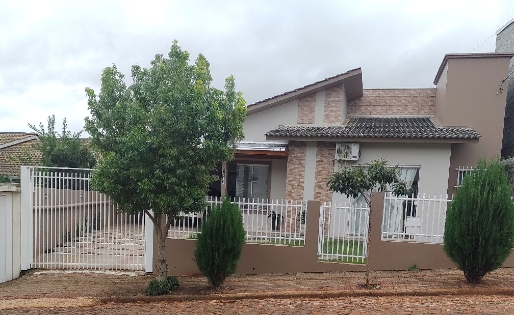 The exterior of a modern home features a combination of brick and stucco siding. A manicured lawn is visible alongside the house, with decorative trees and shrubs positioned by the white picket fence and pathway leading up to the entrance.
