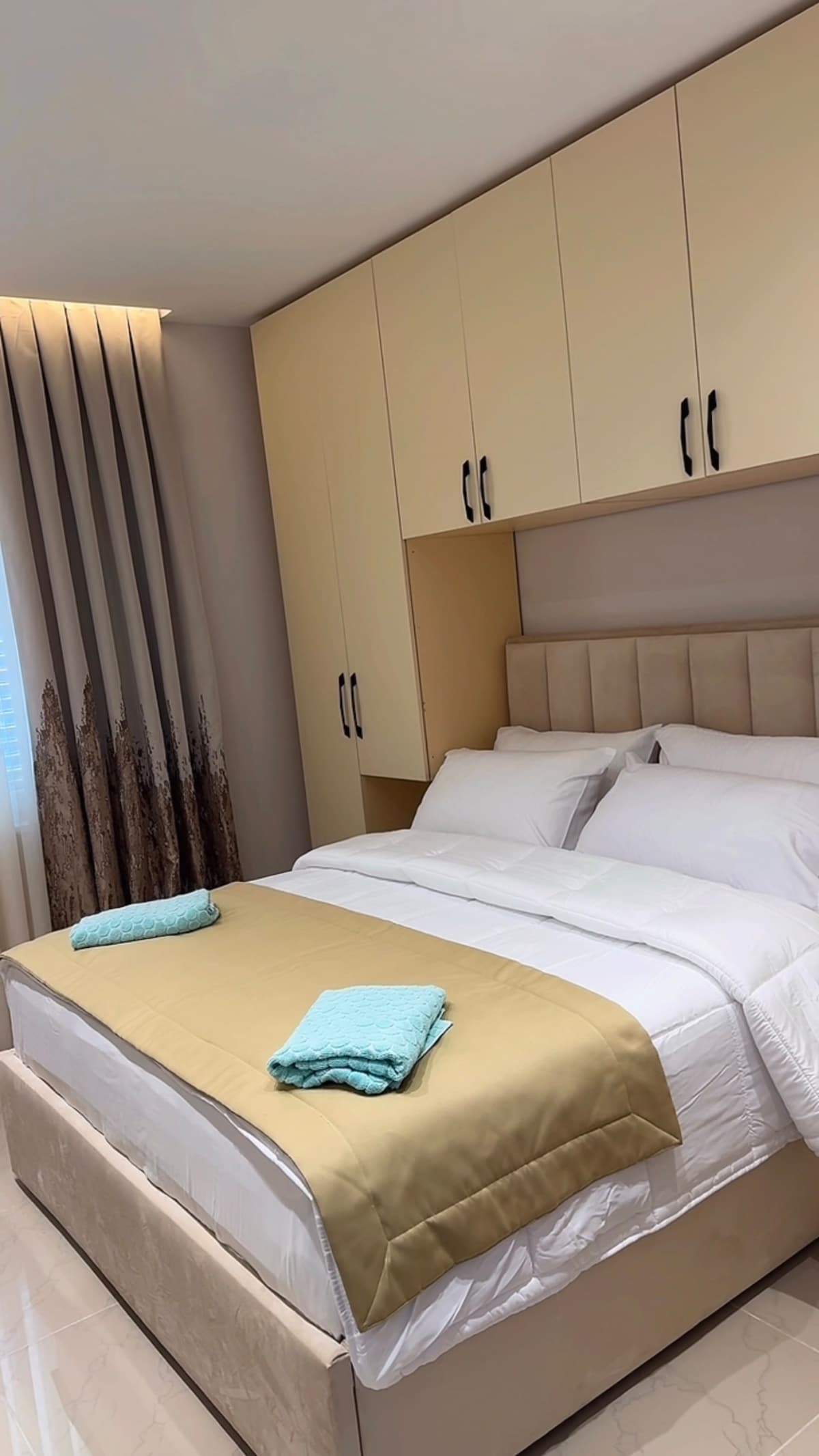 A well-organized bedroom features a spacious bed dressed in crisp white linens and a soft beige throw blanket. Two light blue towels are neatly arranged on the bed. Above, modern cabinets provide ample storage, while light-colored curtains soften the natural light streaming in.