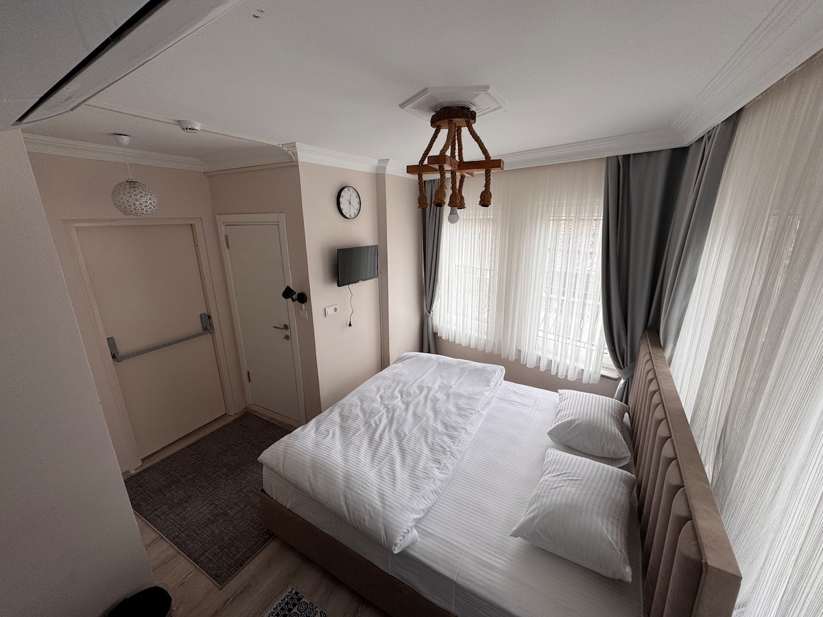 A well-appointed sleeping area features a spacious bed with white linens and two pillows. Natural light filters through sheer curtains, creating a bright and airy environment. Modern decor elements include a wall clock, a mounted television, and a soft area rug enhancing the comfort of the room.
