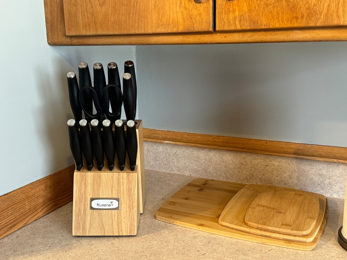 A set of kitchen knives is neatly arranged in a wooden block, featuring black handles. Adjacent to the knife block, a wooden cutting board is positioned on a light-colored countertop with a natural wood grain finish.