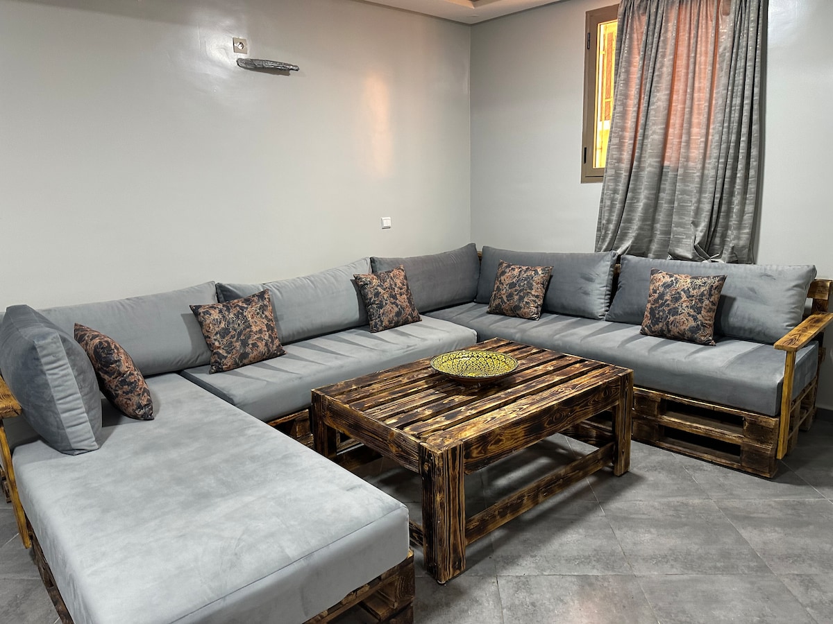 A spacious seating area features a large, L-shaped couch upholstered in soft grey fabric, with decorative cushions in intricate patterns. A rustic wooden coffee table occupies the center, complemented by a round decorative tray. Natural light enters through a nearby window covered with elegant curtains.