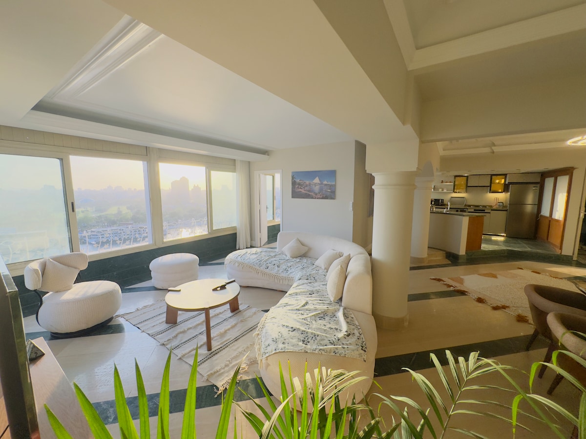 A bright living area features large floor-to-ceiling windows framing expansive views of the Nile. Curved, plush furniture offers a comfortable seating arrangement, complemented by a central coffee table. Subtle indoor greenery enhances the inviting space, which is infused with natural light.