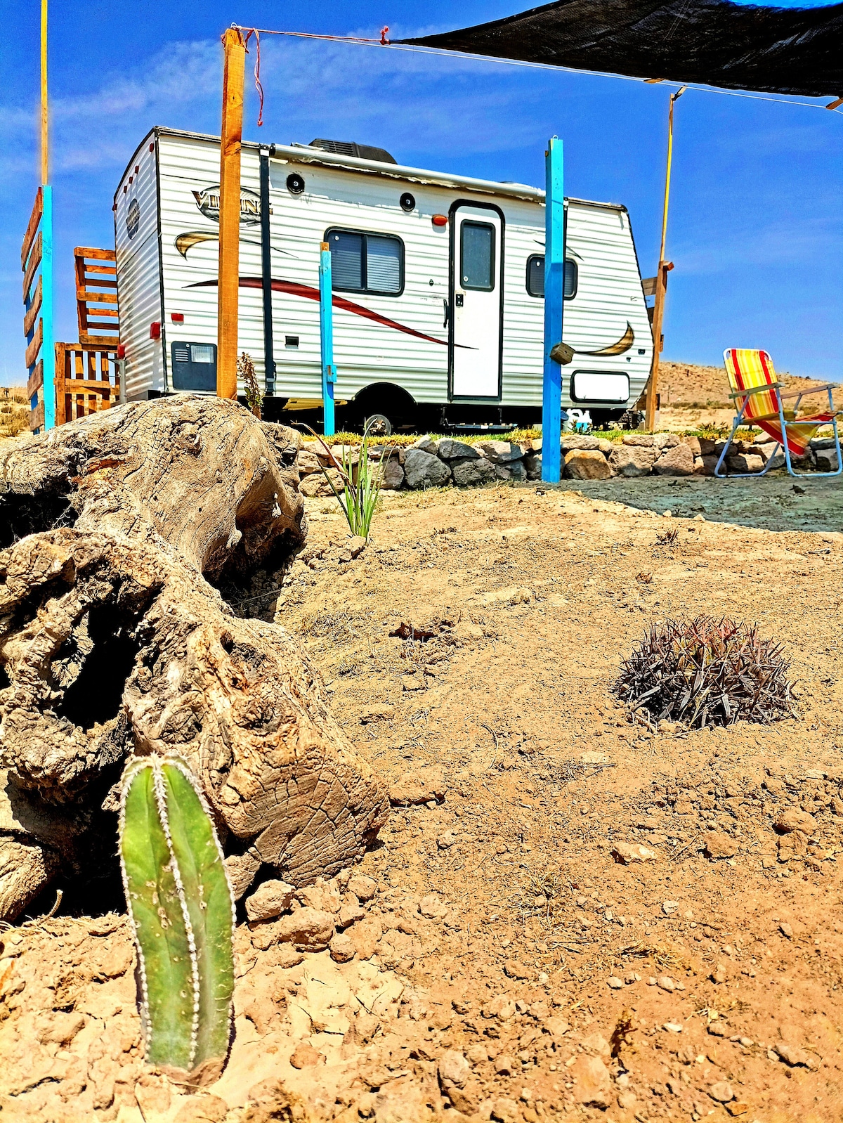A cozy camper is positioned in a desert landscape, surrounded by arid terrain and native plants. Sunlight brightens the scene, with a colorful chair and a shaded area visible in the background. Rock formations and cacti add natural accents to the outdoor space.