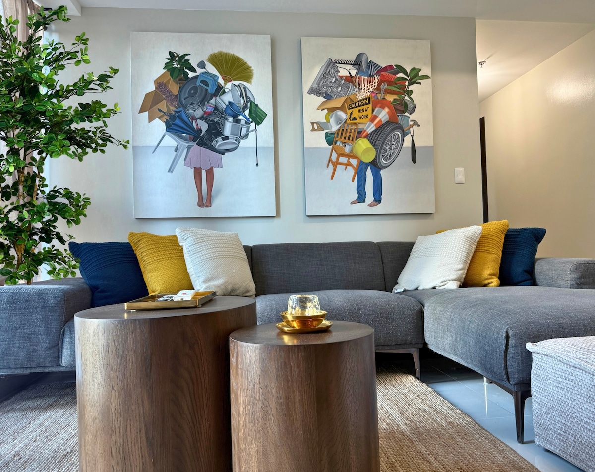 A spacious living area features a comfortable gray sofa adorned with patterned pillows in yellow and white. Two wooden coffee tables sit in front of the sofa, complemented by a decorative tray. Art pieces depicting figures and various objects are hung on the walls.