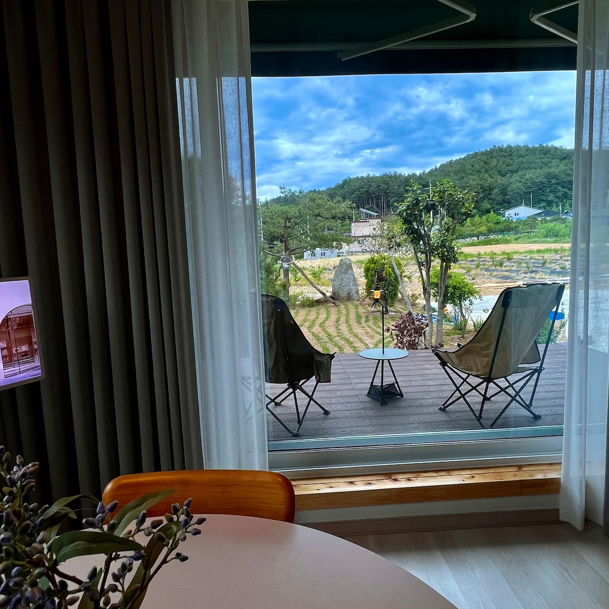 A view from the indoor living space features two comfortable chairs positioned on a wooden deck. Lush greenery and garden elements are visible beyond, framed by sheer curtains that softly diffuse natural light. The overall scene reflects a serene outdoor atmosphere.
