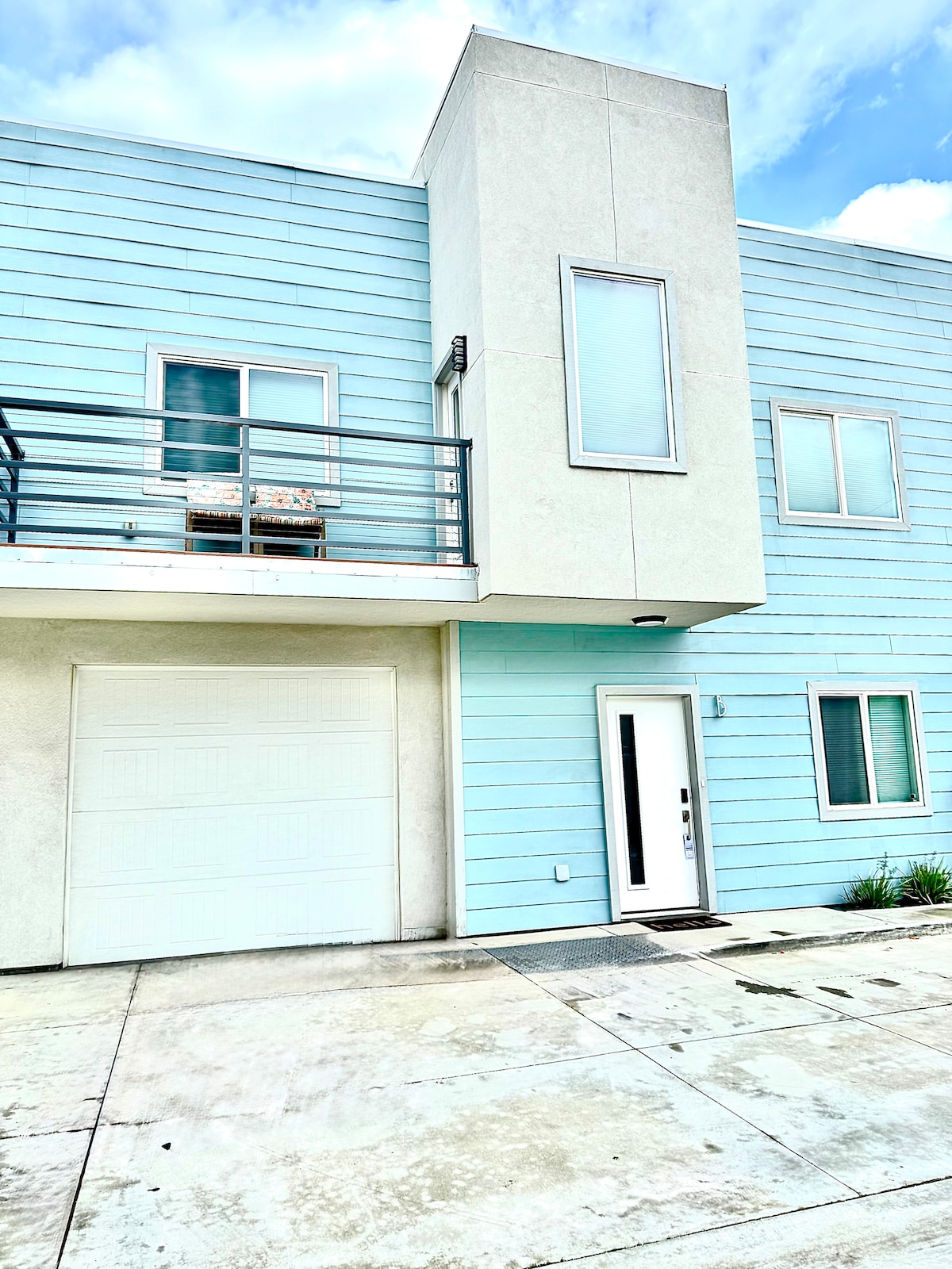 The exterior of a modern, two-story home showcases a light blue facade with a mix of large windows and a balcony. A private garage door is positioned on the right, while the front entrance features a sleek door. A small patch of greenery is visible along the base.