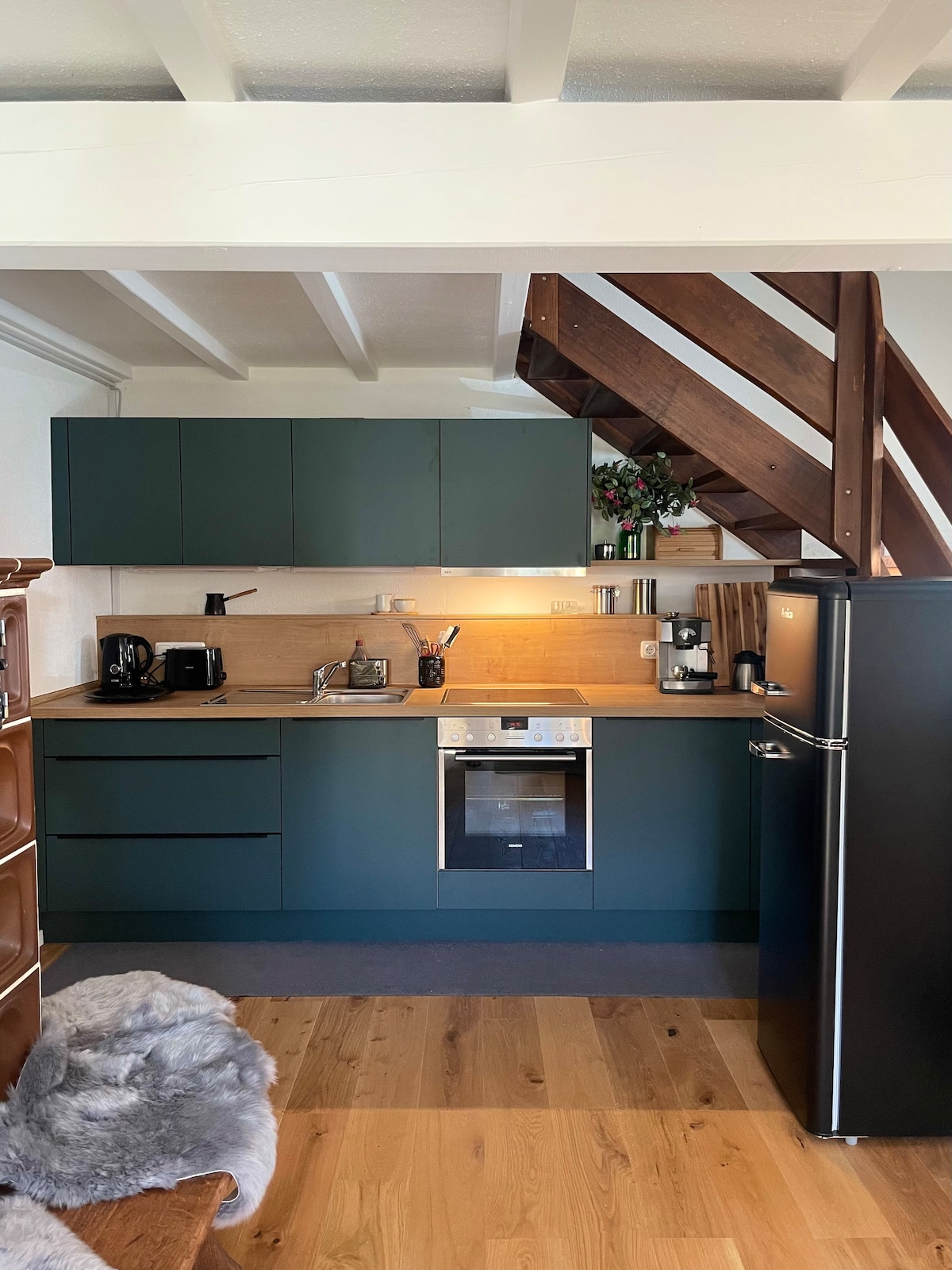 A modern kitchen setup is presented, featuring dark green cabinetry and wooden accents. Appliances include a refrigerator, oven, and coffee maker, contributing to a well-equipped space. Natural light enhances the warm wood flooring, while decorative plants add a touch of greenery.