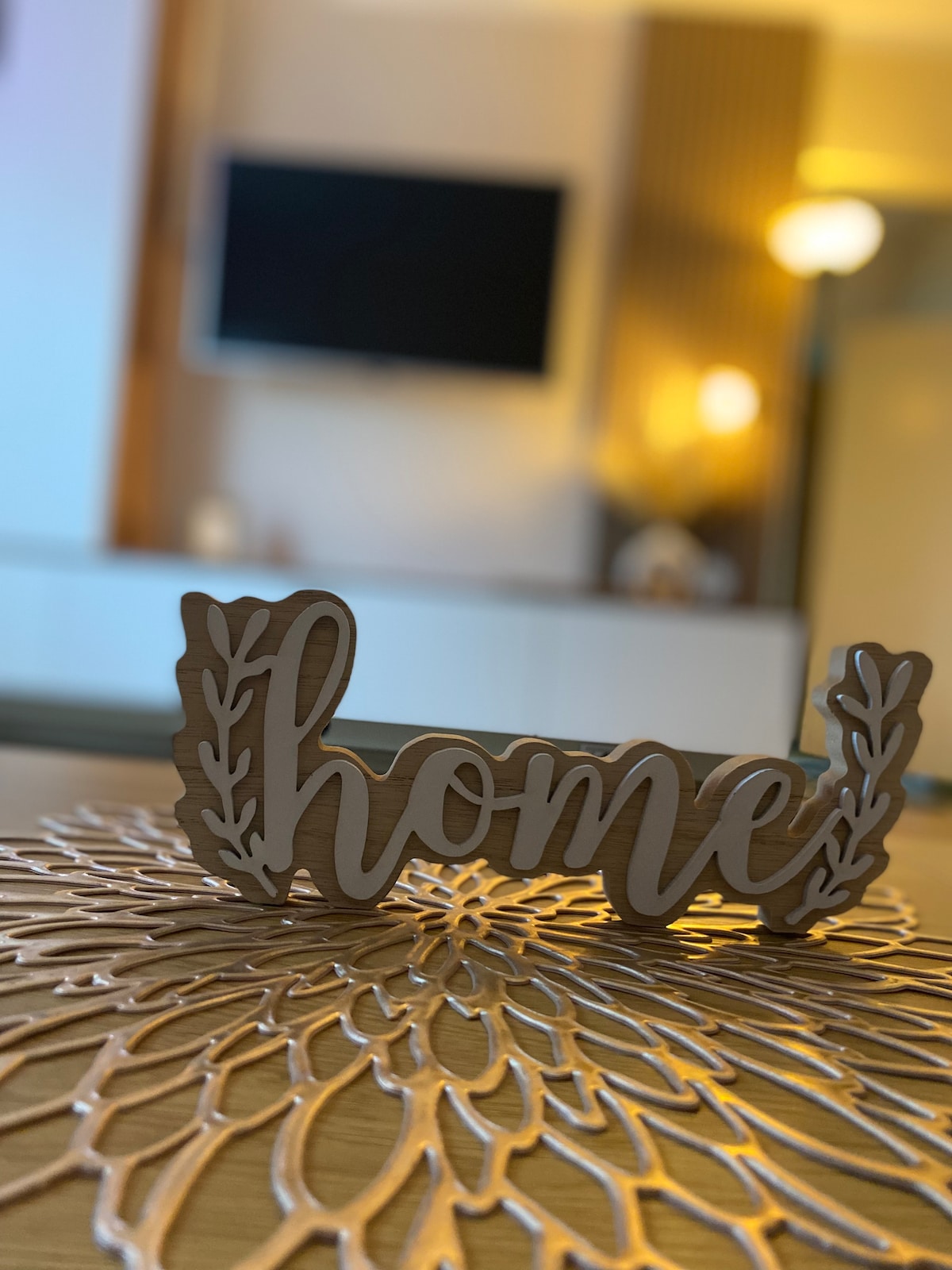 A decorative wooden sign reading 'home' is positioned on a detailed, lace-like placemat. In the background, a softly illuminated television stands against a tranquil wall, contributing to a cozy ambiance in the living space.