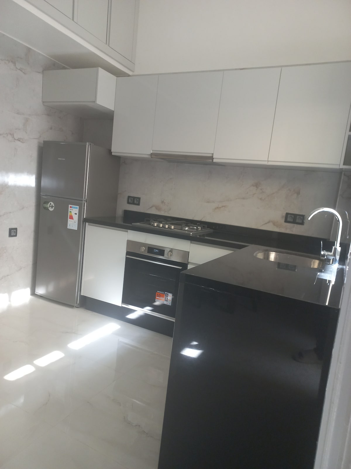 A modern kitchen features sleek black countertops and white cabinetry. Stainless steel appliances, including a refrigerator and stovetop, complement the clean design. Bright light illuminates the space through large windows, enhancing the contemporary feel of the kitchen.