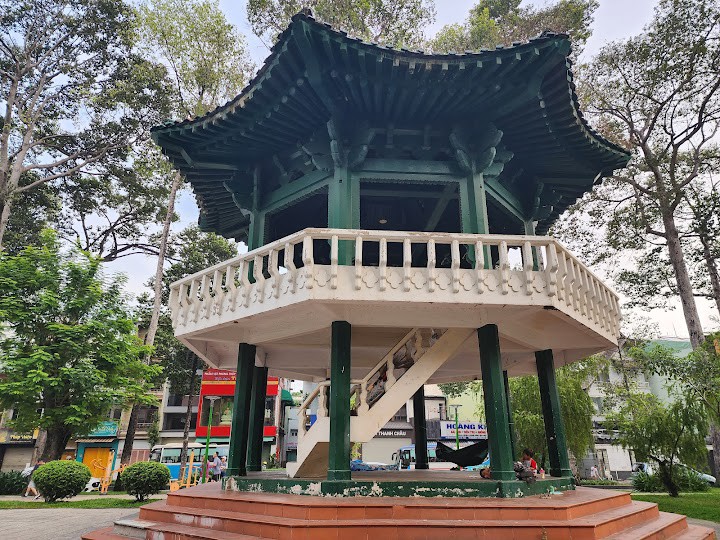 A traditional-style pavilion is situated in a park, with a circular base and wooden details in dark green. A wide staircase leads up to the open area, surrounded by a white railing. Trees provide shade around the structure, enhancing the tranquil setting.