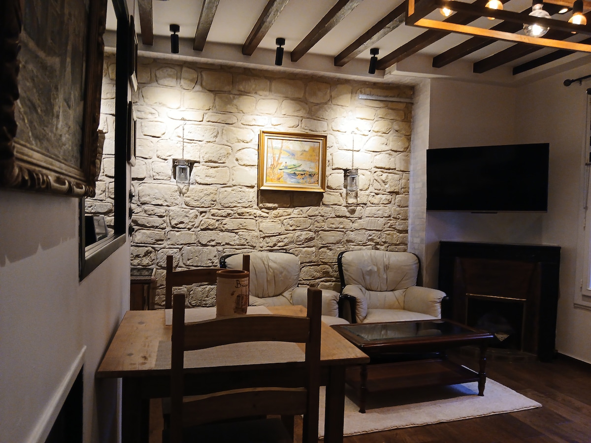 A living area features exposed wooden beams and a stone wall with soft lighting. A painting is hung above a cozy seating arrangement, which includes two armchairs. A wooden dining table is positioned nearby, with a glass top coffee table in front of the seating.