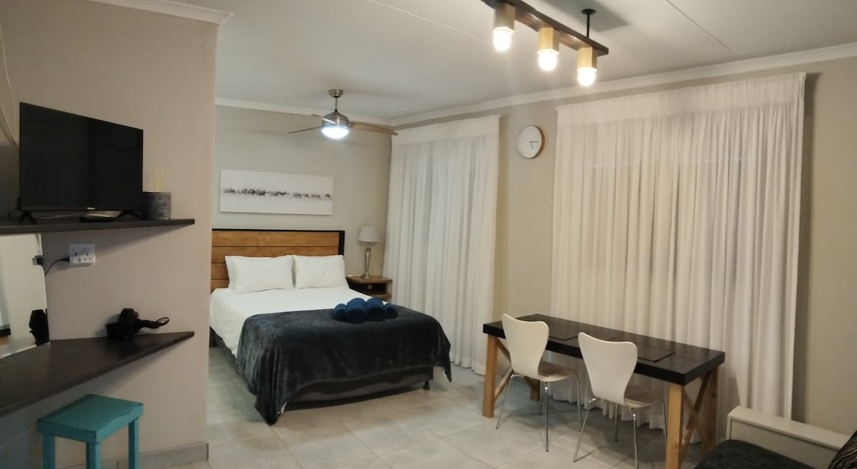 A well-lit studio space features a spacious bed with a dark blue throw and two decorative pillows. A dining table with four white chairs is visible. Bright, sheer curtains partially cover a window, and a TV is mounted on a wall shelf.