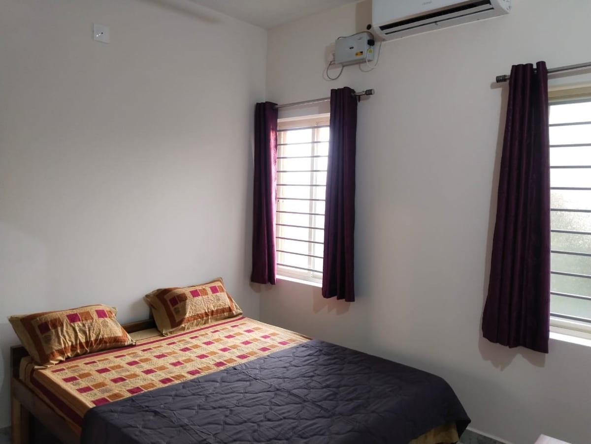 The bedroom features a spacious bed with a patterned bedspread and contrasting pillows. Two large windows, dressed with dark curtains, allow natural light to filter in. The walls are painted in a soft tone, and an air conditioning unit is mounted above.