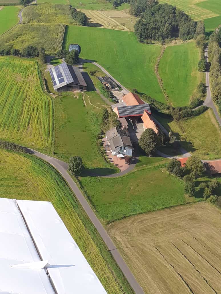 An aerial view captures a cluster of buildings set in lush green fields. Roads wind through the landscape, surrounding the structures, some with red roofs. Nearby, fields of varying shades of green and brown stretch out, providing a serene rural backdrop.