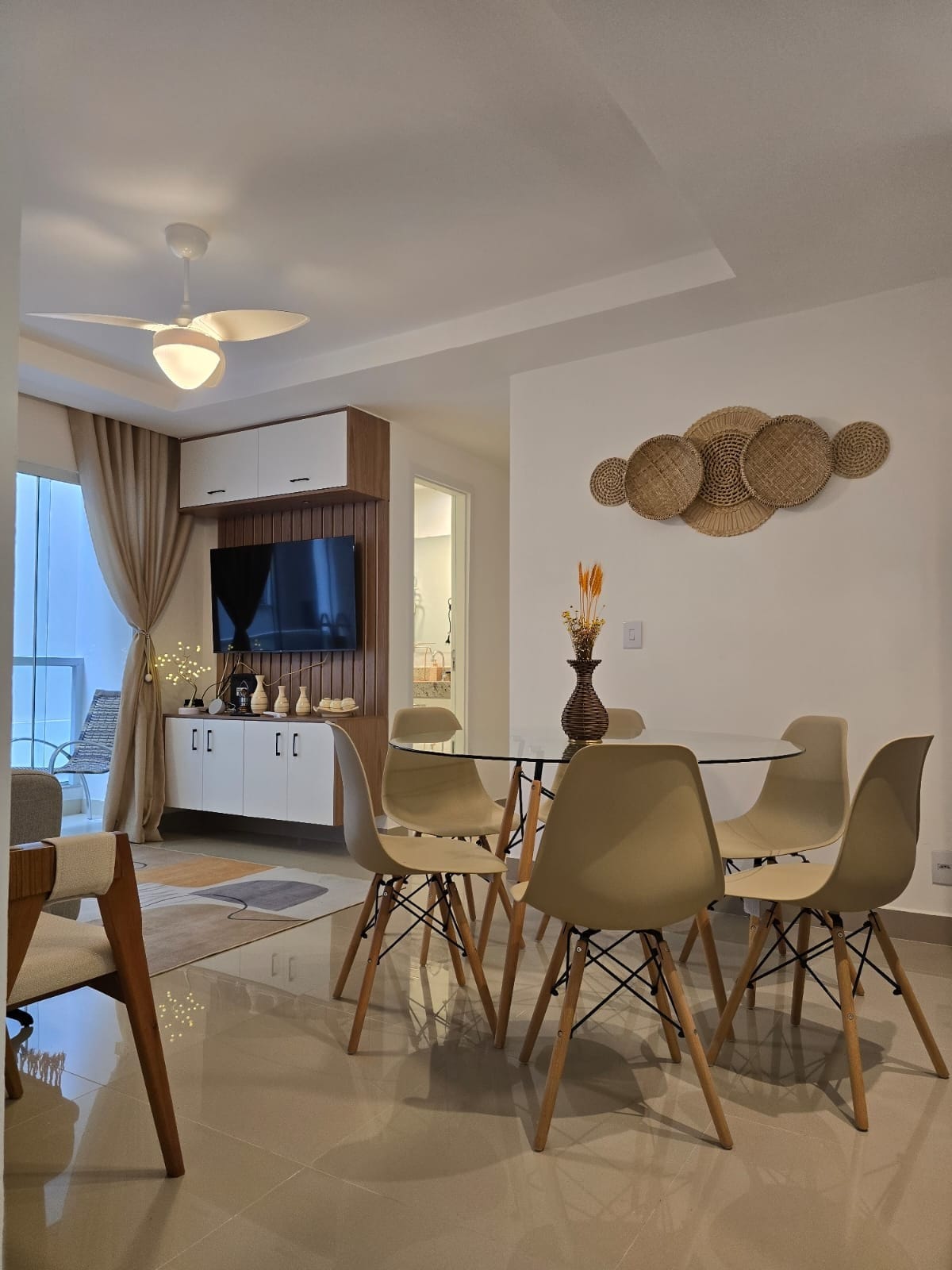 A modern dining area is presented with a round glass table surrounded by six light-colored chairs. A decorative wall feature composed of woven materials adds texture. Soft lighting is provided by a ceiling fan, creating a comfortable ambiance.