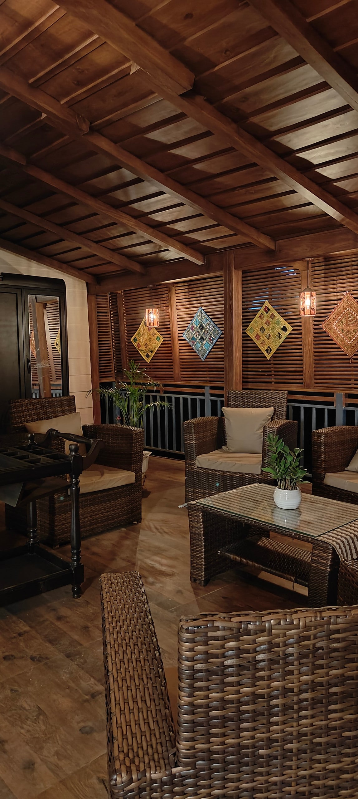 A cozy seating area features woven rattan chairs surrounding a small table, set in a wooden gazebo. The space is brightly illuminated by wall sconces and showcases colorful wall hangings. Slatted wooden walls offer a sense of privacy while allowing natural light to filter in.