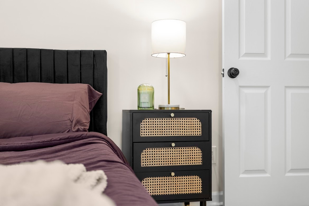 A cozy sleeping area features a dark upholstered headboard with plush bedding in a rich hue. A stylish nightstand with cane detail sits beside the bed, topped with a modern lamp and a decorative candle. A white door is visible in the background.