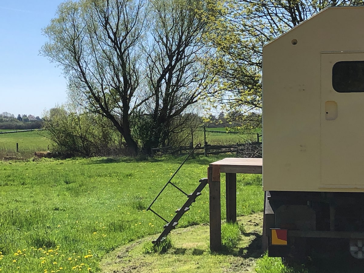 A rustic trailer stands adjacent to a grassy area, with a small wooden deck featuring steps leading up. A large tree and open fields are visible in the background, complemented by a clear sky and natural greenery.