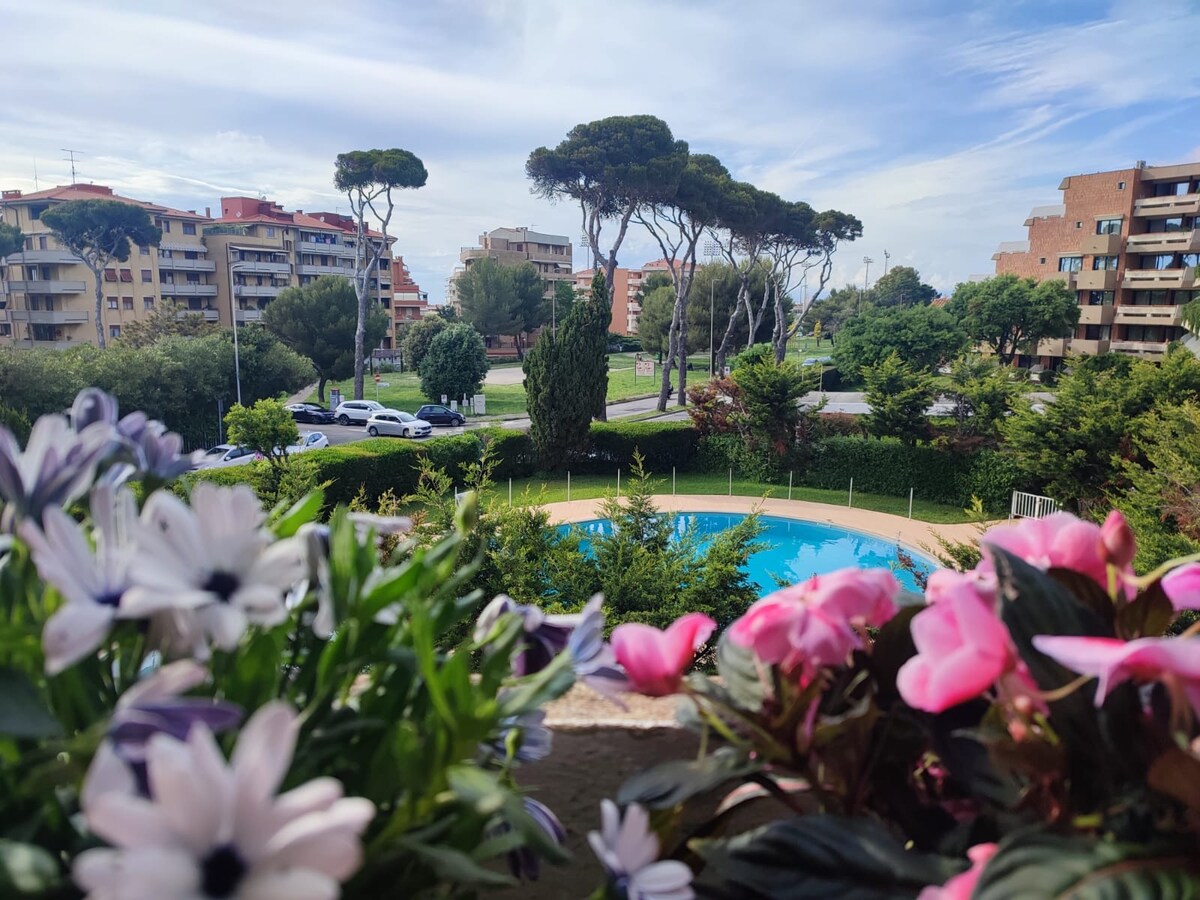 The image presents a view from a balcony, featuring vibrant flowers in the foreground. A clear pool is visible among lush greenery and trees, with buildings in the background. The serene colors and natural elements contribute to a calming atmosphere.