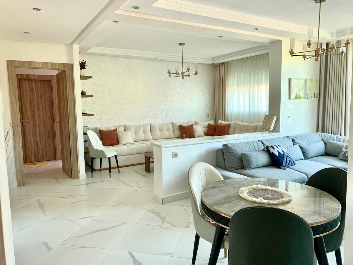 A spacious living area features light-colored walls and a large sectional sofa adorned with accent pillows. A round dining table with dark chairs is positioned nearby. Natural light filters through sheer curtains, enhancing the modern decor.