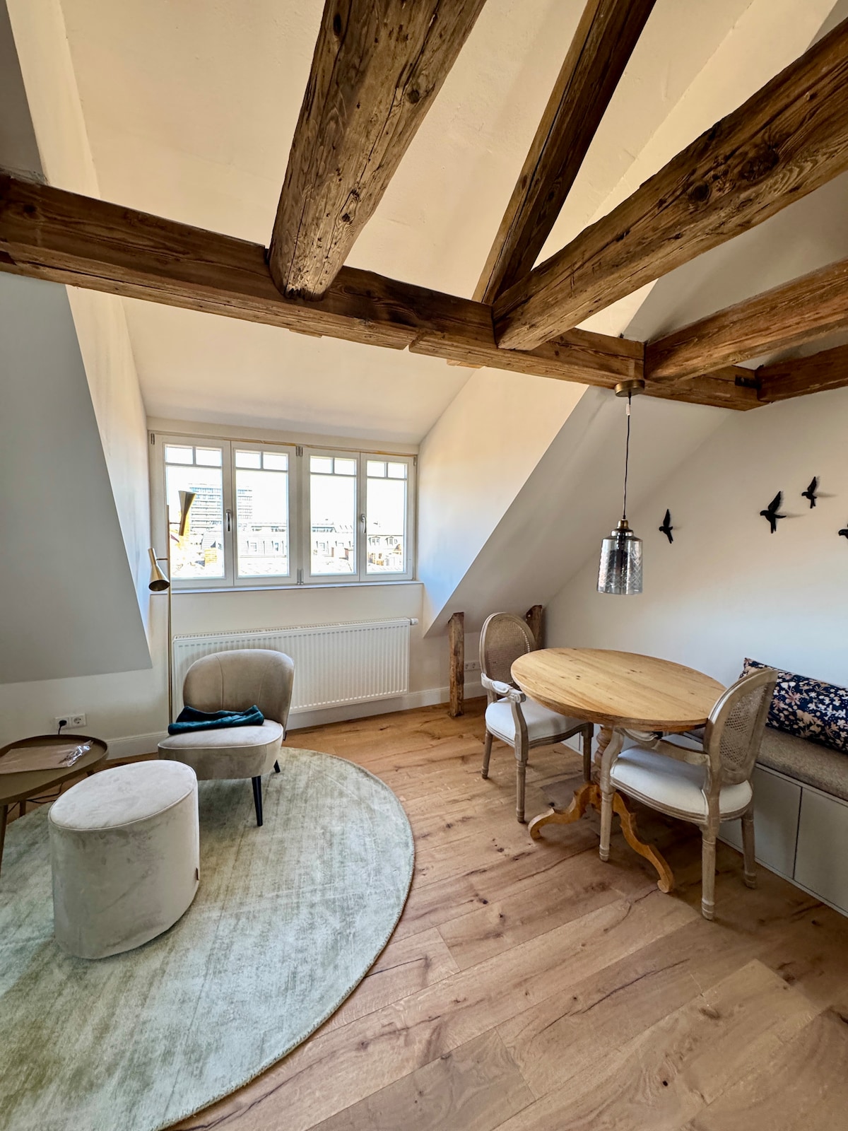 A bright living space is showcased, featuring exposed wooden beams and a circular area rug. A small, round dining table with chairs is positioned near a large window, providing natural light. A cushioned bench and decorative accents add to the inviting ambiance.