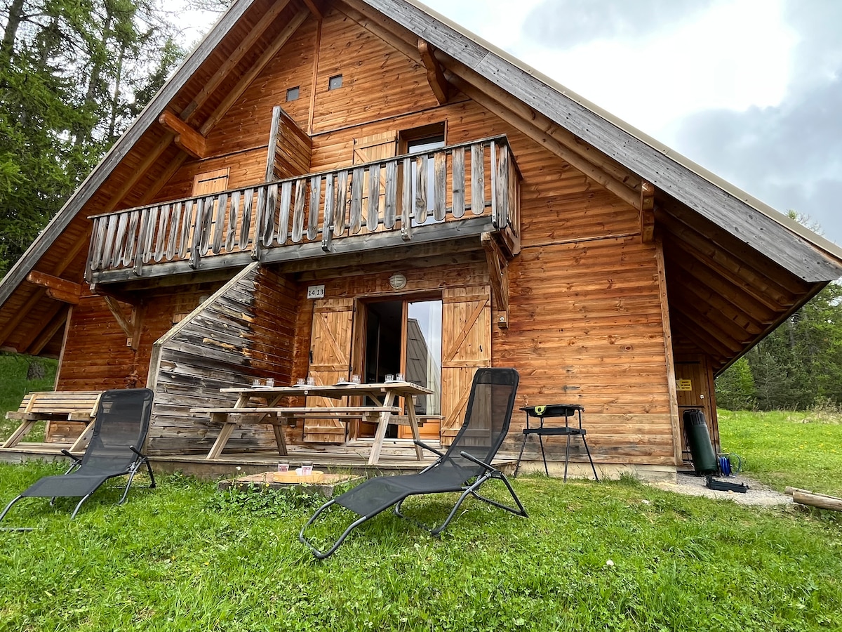 The wooden chalet features a well-maintained exterior with a spacious balcony and steps leading to the entrance. Two lounge chairs sit on the grassy area in front, promoting relaxation. The surrounding trees offer a serene natural backdrop.