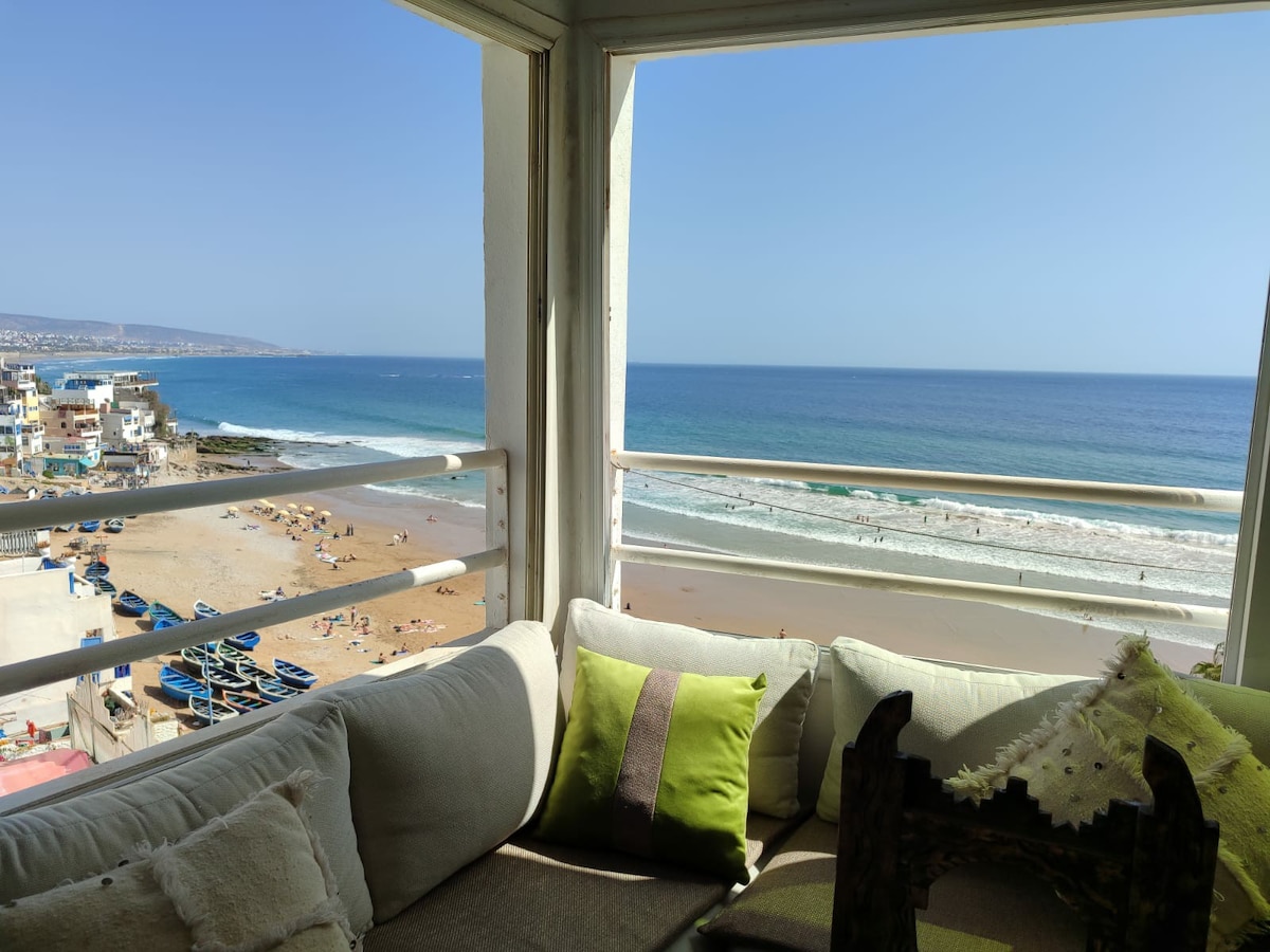 A cozy seating area is positioned in a bright corner, featuring plush pillows of varying shades and a view of the ocean through large windows. The beach below is visible, with a sandy shore and gentle waves, alongside beachgoers and colorful fishing boats.