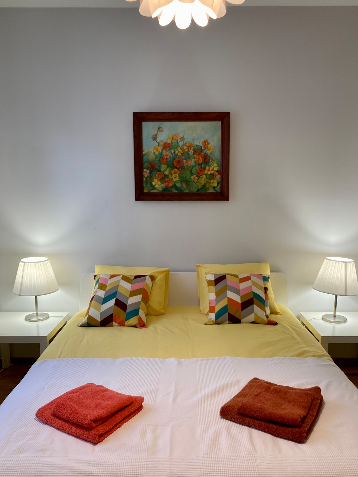 A cozy bedroom is furnished with a spacious bed adorned with colorful pillows and blankets. Two bedside lamps provide soft lighting, while a floral painting adds a touch of warmth to the decor. The overall setup encourages a restful atmosphere.