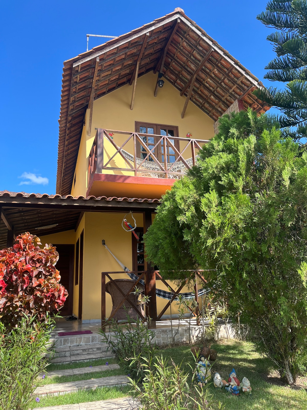 A charming yellow house with a sloped roof is framed by lush greenery and flowering shrubs. A balcony is visible on the upper level, and a welcoming entrance leads to the path. Garden ornaments add character to the outdoor space.