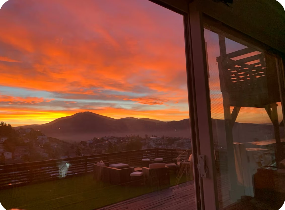 A breathtaking sunset view showcases vibrant orange and pink hues filling the sky above rolling hills. The scene includes an outdoor deck area with a dining table and chairs, framed by expansive glass doors, inviting the beauty of nature indoors.