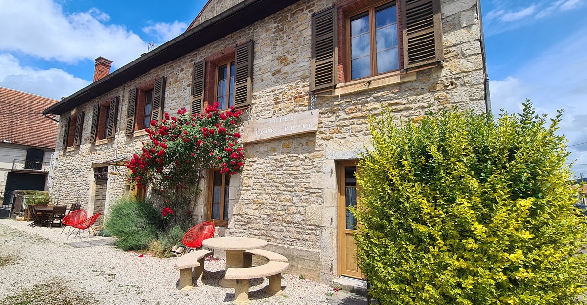 The stone facade of the house is complemented by bright red flowers and greenery surrounding the entrance. A circular stone seating area and modern red chairs create an inviting outdoor space. Large windows with wooden shutters enhance the home's rustic charm.