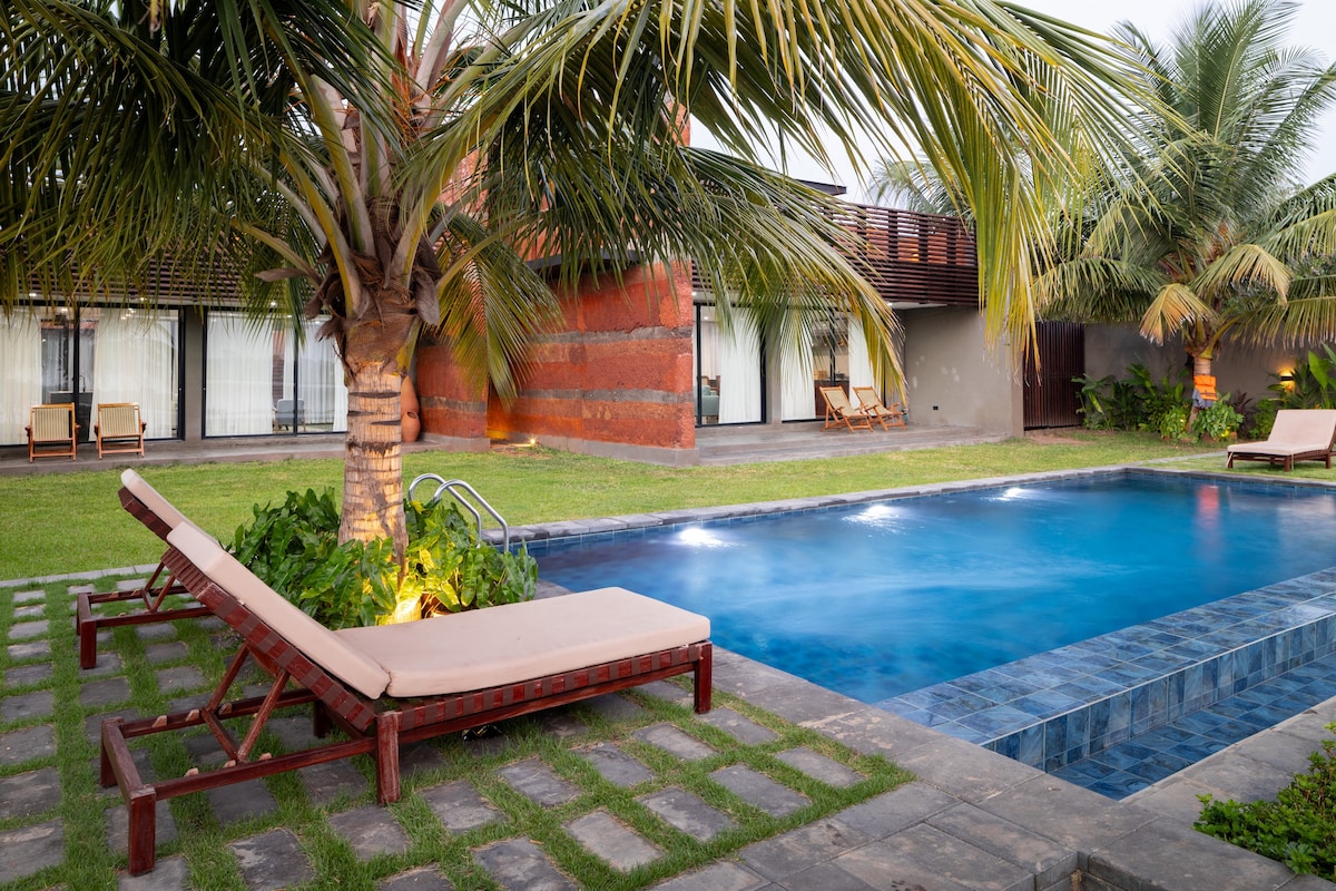 A serene outdoor space features a swimming pool surrounded by tropical palm trees. Lounge chairs are positioned beside the pool, while well-maintained grass and stone pathways enhance the relaxing ambiance. The building's large windows reflect natural light, adding to the inviting atmosphere.