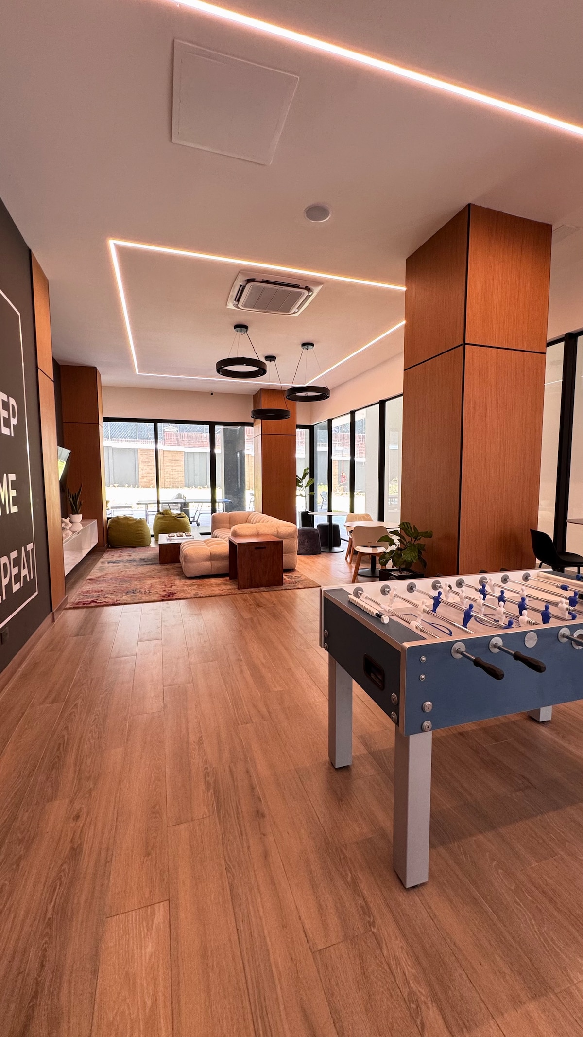 The communal area features modern architectural elements with wooden accents. A foosball table is positioned in the foreground, while the background includes a cozy seating arrangement, natural light flowing through large windows, and a warm ambiance created by stylish light fixtures.