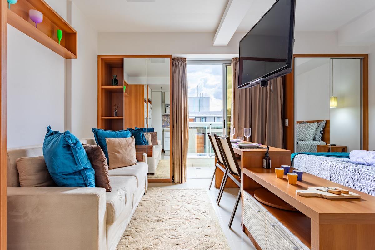 A modern living area is presented with a comfortable sofa adorned with colorful cushions. A wooden coffee table sits in front, while a wall-mounted television is visible. Mirrors reflect light and space, enhancing the openness of the room, which connects to a balcony with natural light.