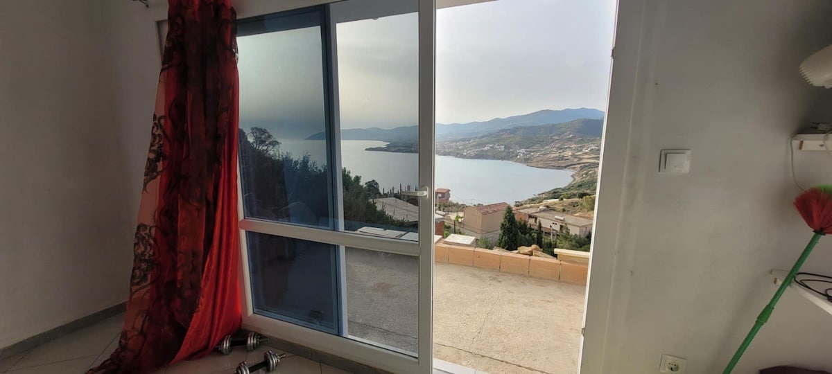 A large window reveals a scenic view of the coastline and mountains beyond. A portion of the room is visible, featuring light-colored walls and tiled flooring. Sheer curtains frame the window, allowing natural light to fill the space.